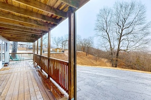 Une terrasse en bois avec un banc, une rambarde et une vue sur un paysage aride, parsemé d’arbres et de collines.