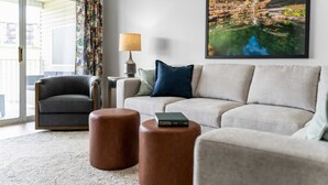 Een woonkamer met een bank, fauteuil, twee poefjes, een lamp en een ingelijst schilderij aan de muur.
