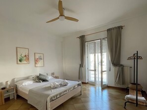 A bedroom with a bed, a bedside table, a nightstand, a ceiling fan, a window with curtains, and a radiator. - Ostuni Home with Terrace & Free Parking – Near Old Town (Ostuni)