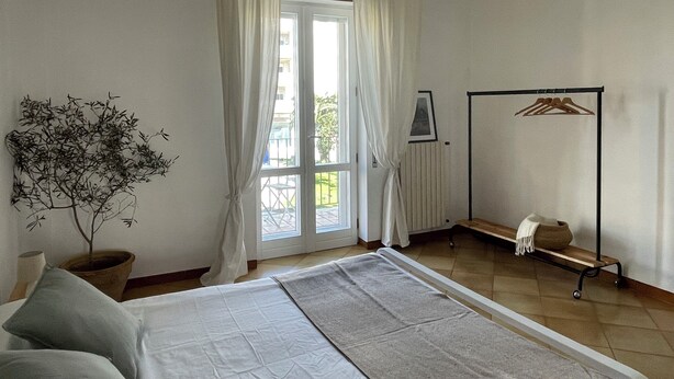 Una camera da letto con un letto, una panca, un termosifone, una pianta e una finestra con tende.