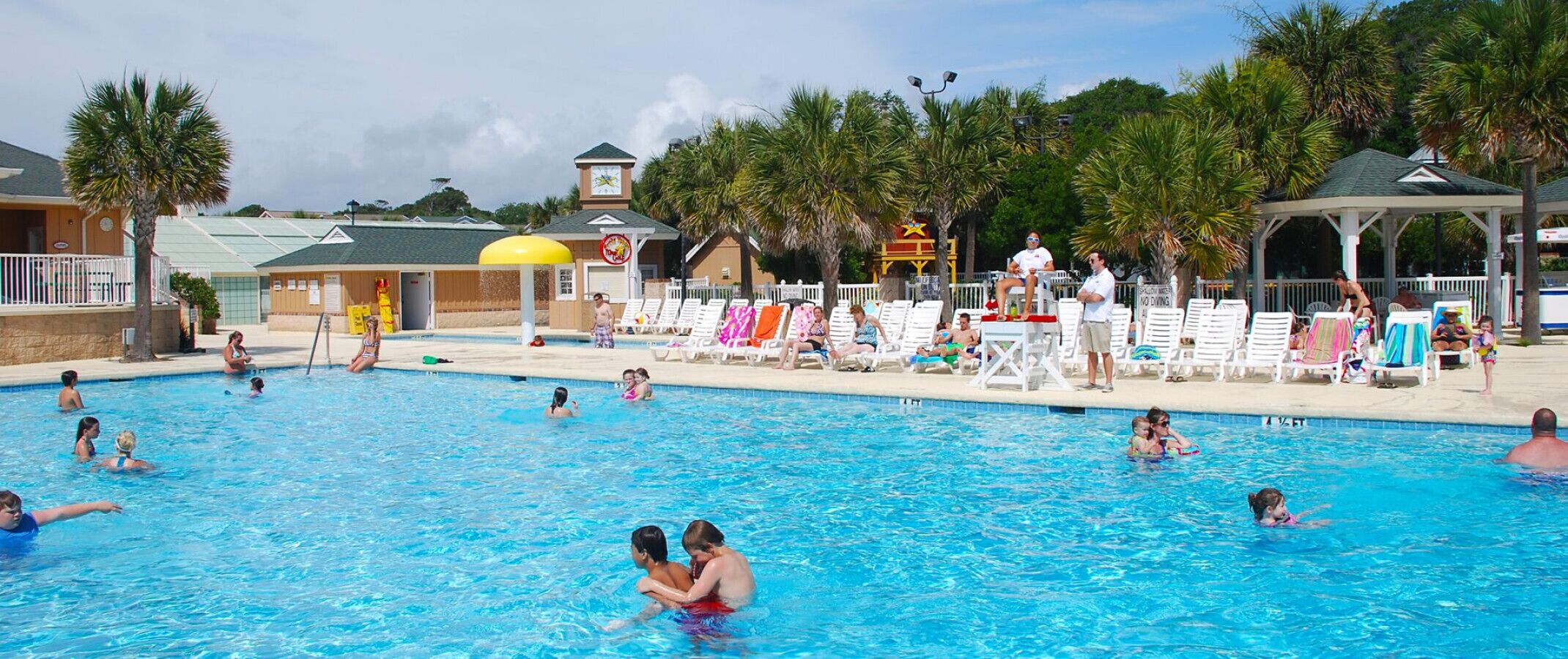 A swimming pool with people in the water and on the deck, surrounded by palm trees and a building with a sign.