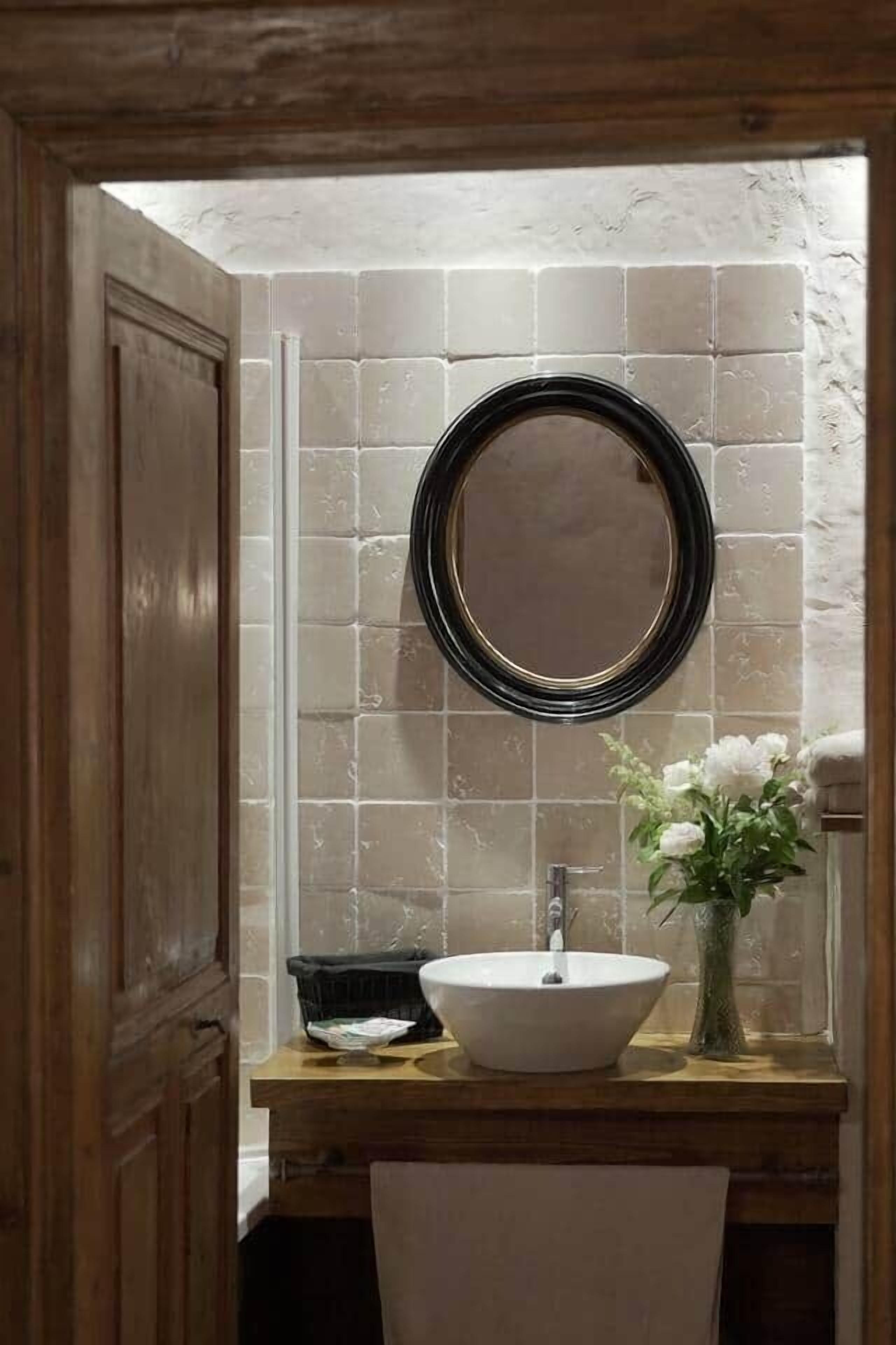 A bathroom with a wooden vanity, a white sink, a round mirror, and a vase with flowers.