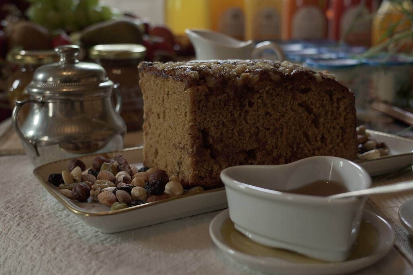 Eine Kuchenscheibe mit Nüssen auf einem Teller, eine kleine Schale mit Sirup und ein silbernes Teekännchen.