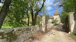 Un chemin de pierres mène à un bâtiment en pierre surmonté d’un toit conique, le tout étant entouré d’arbres et de verdure.