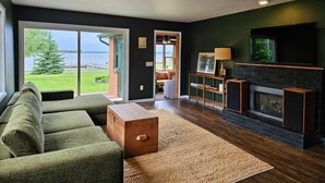 A living room with a large window, a fireplace, and a view of a lake. - Hessel Bay Main Cabin : Idyllic Les Cheneaux Islands waterfront cabin sleeps 8 (Hessel)