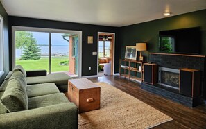 A living room with a large window, a fireplace, and a view of a lake. - Hessel Bay Main Cabin : Idyllic Les Cheneaux Islands waterfront cabin sleeps 8 (Hessel)