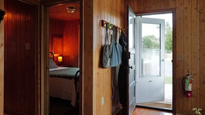 A wooden interior with a bed, a door leading outside, and a coat rack with bags. - Hessel Bay Cabin 1:  Lake Huron waterfront resort cabin sleeps 2. (Hessel)