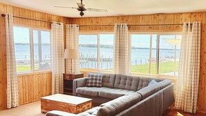 A cozy living room with a sectional sofa, a small table, and a lamp. Large windows offer a view of the water and dock. - Hessel Bay Cabin 1:  Lake Huron waterfront resort cabin sleeps 2. (Hessel)