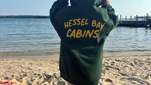 A person wearing a green hoodie with "Hessel Bay Cabins" written on the back, standing on a sandy beach with water in the background. - Hessel Bay Cabin 2:  Lake Huron waterfront resort cabin sleeps 2. (Hessel)