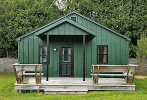 A green wooden cabin with a front door, two windows, and a porch with benches.