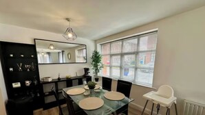 A modern dining area with a glass table, black chairs, a white high chair, a black console, and a large window with blinds. - 2br city side apartment (Belfast)