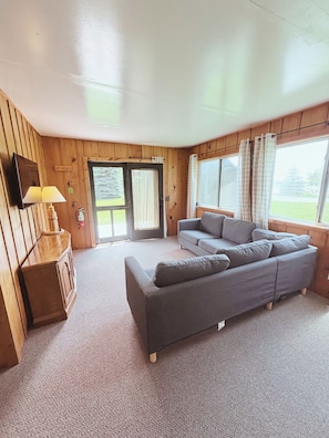 A room with a grey sofa, a wooden cabinet, a lamp, and a large window with curtains.