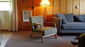 Une pièce avec un mur lambrissé en bois, un fauteuil vert, un canapé gris, un lampadaire, une horloge murale et une fenêtre avec des rideaux.