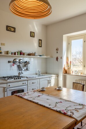 Uma cozinha com fogão a gás, armários brancos, uma mesa de madeira com toalha de mesa florida e um lustre.