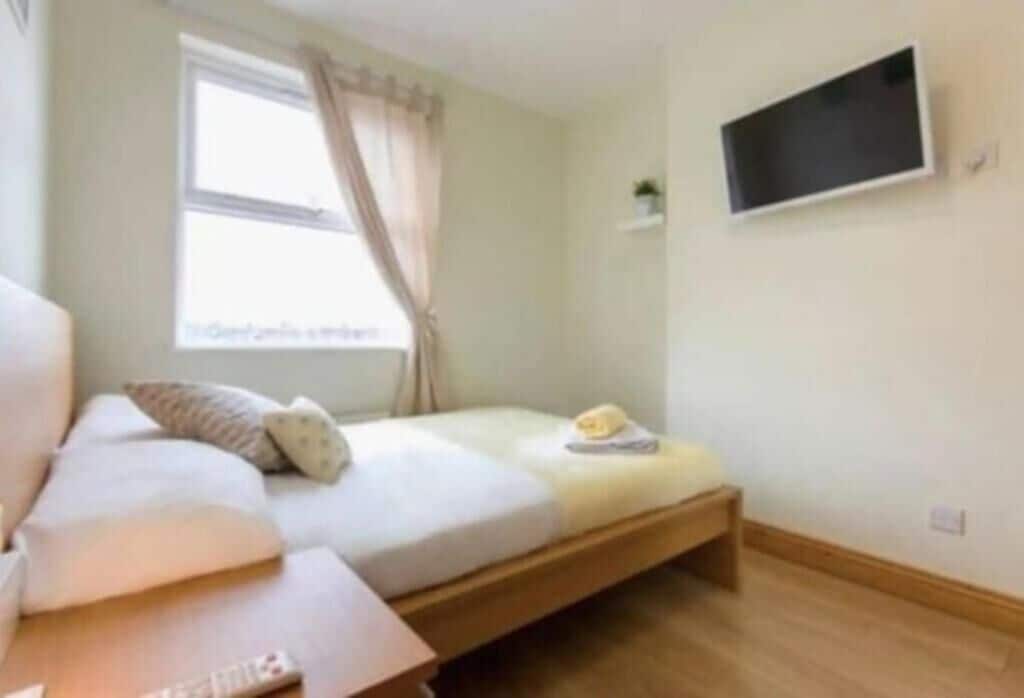 Une pièce comprenant un lit, un téléviseur fixé au mur, une fenêtre avec des rideaux et une petite étagère.