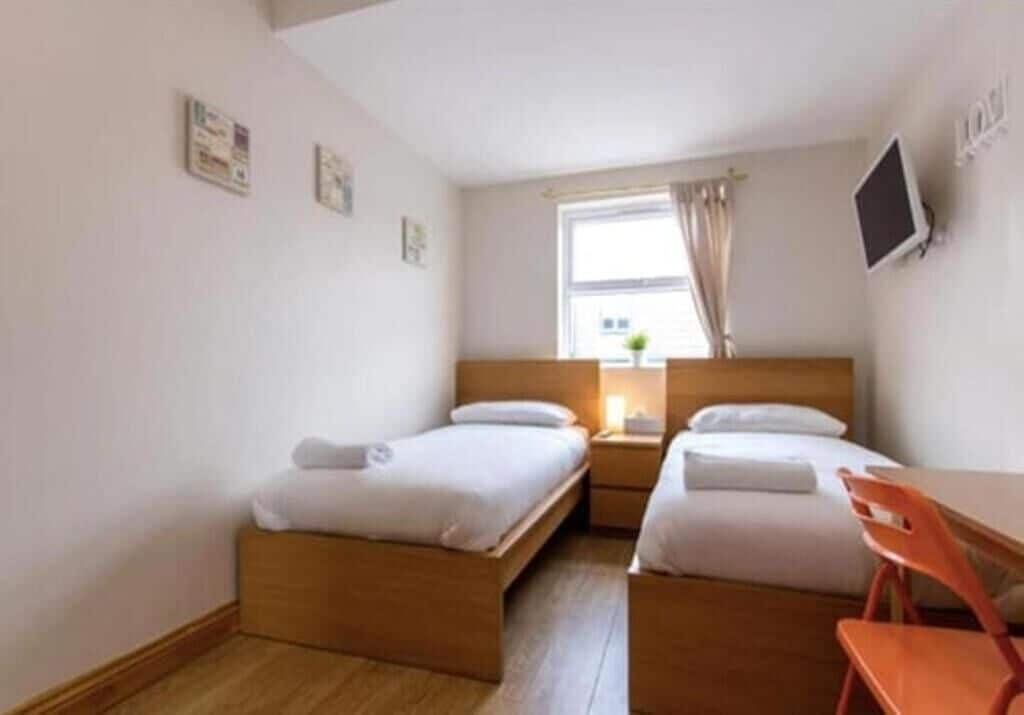 A small, clean room with two single beds, a wooden desk, a chair, a TV, and a window with curtains.
