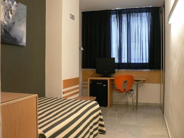 Une petite chambre d’hôtel avec un lit, un bureau avec un ordinateur, une chaise et une fenêtre avec des rideaux.