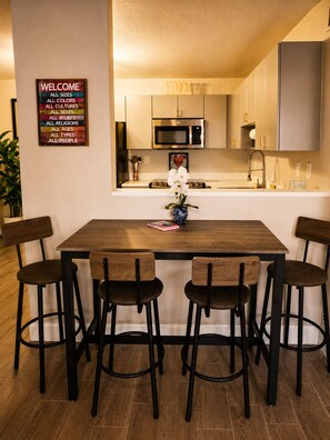 A modern kitchen with stainless steel appliances, a wooden dining table with bar stools, and a welcome sign on the wall.