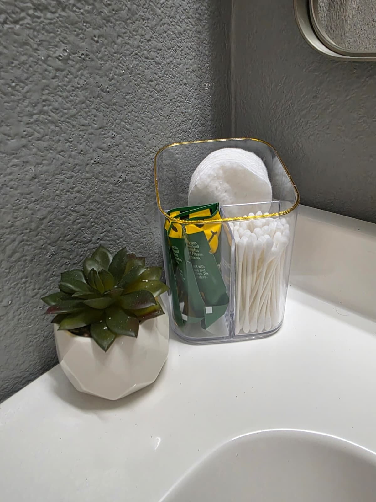 Un lavamanos con un recipiente de vidrio transparente que contiene hisopos de algodón y un paquete verde, junto a una pequeña planta suculenta en maceta.
