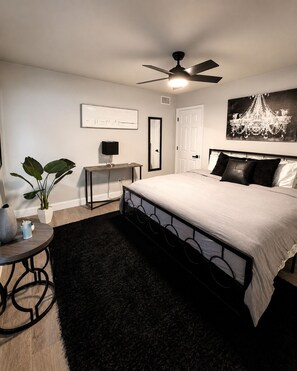 A bedroom with a bed, a ceiling fan, a desk, a chair, a plant, and a chandelier.