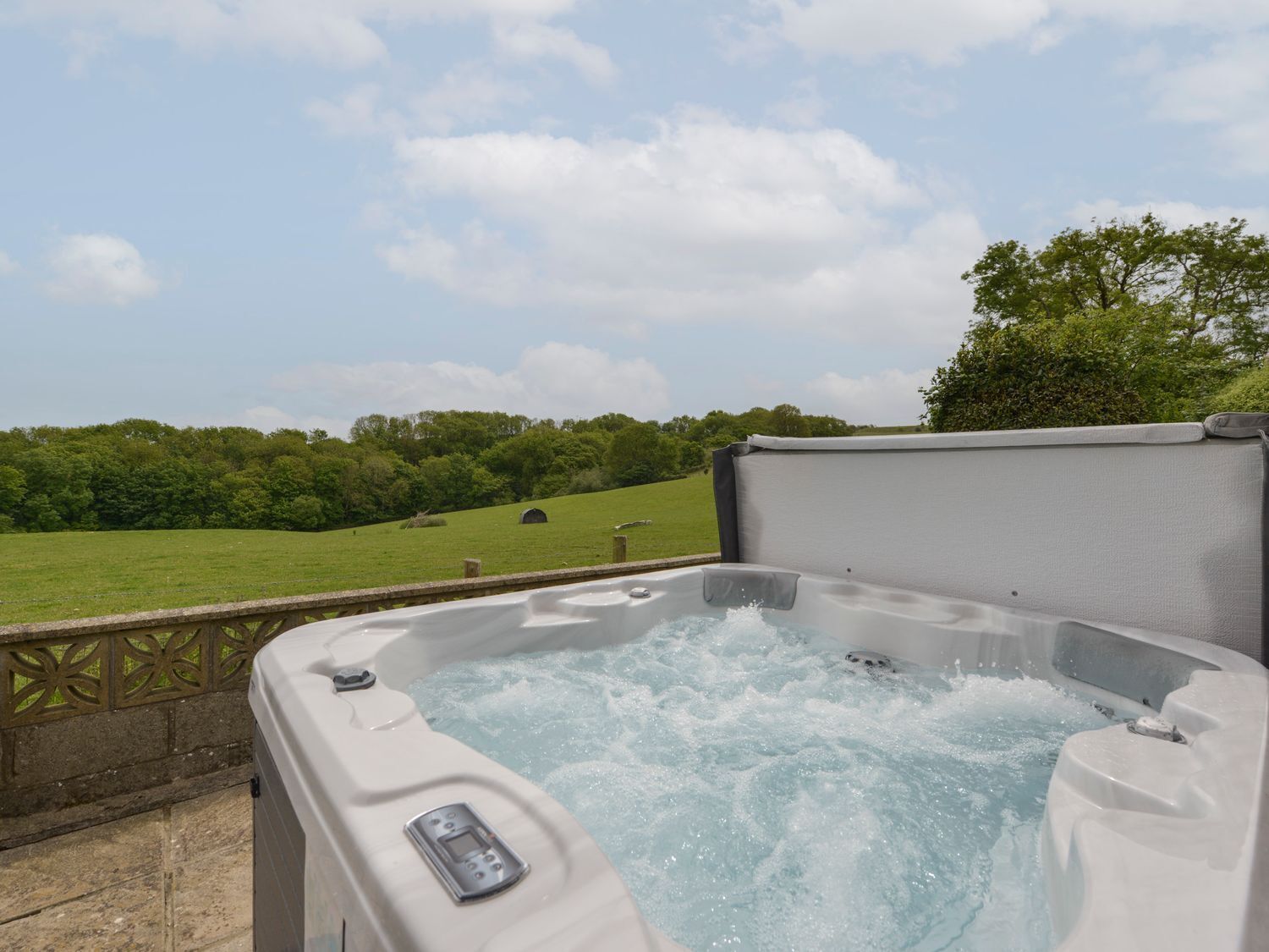 A hot tub with a control panel, surrounded by a stone wall and a view of a green field and trees.