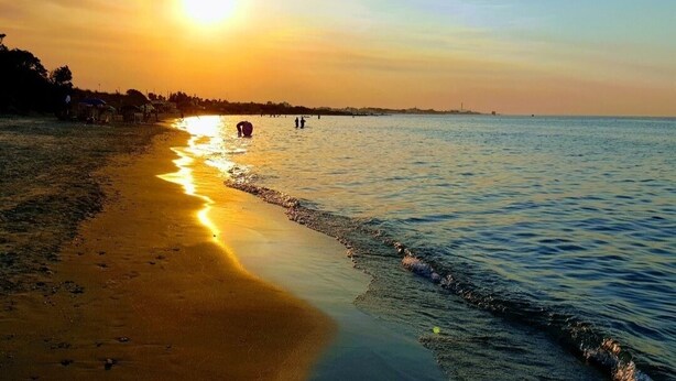 Une scène de plage au coucher du soleil, avec une eau calme et quelques personnes au loin.