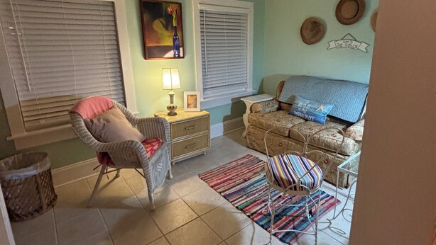 Una habitación con ventilador de techo, dos ventanas con persianas, una silla de mimbre, una mesita con lámpara, un sofá y una alfombra colorida.