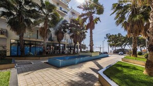 A tiled walkway with a rectangular pool, palm trees, and a building with balconies. - Beachfront Apartment with Pool Access by PikHost (Shëngjin)