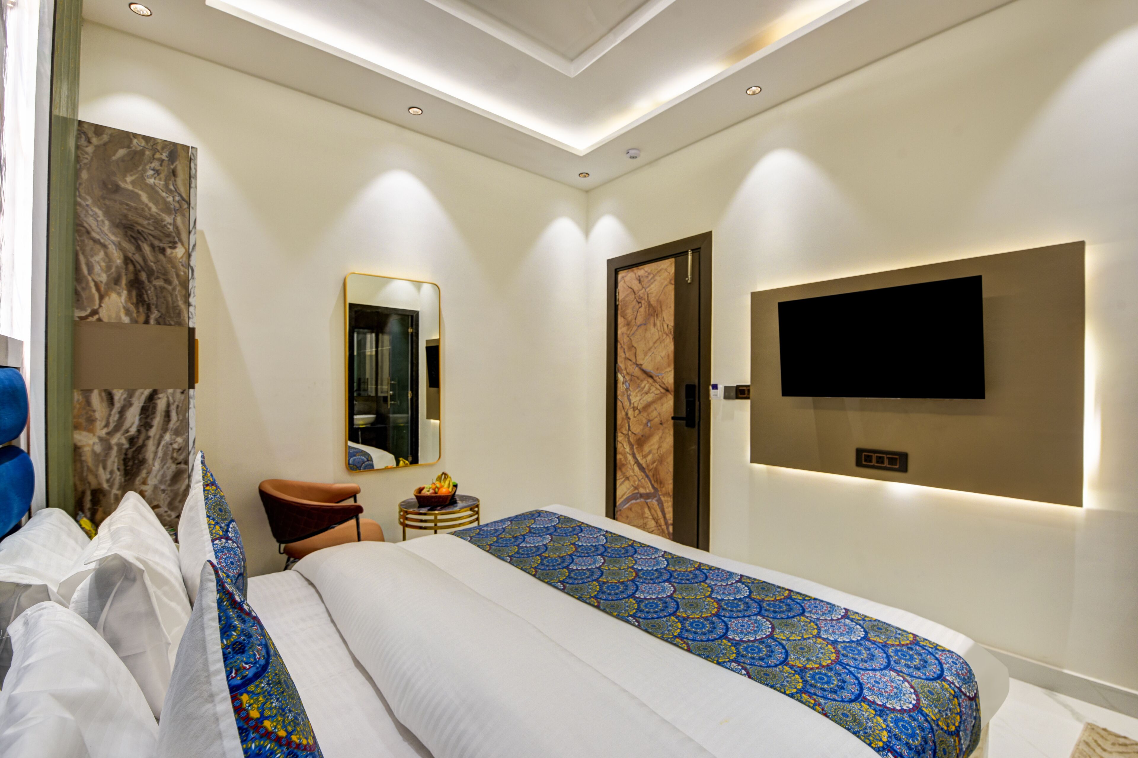 Una habitación de hotel moderna con una cama grande, un televisor de pantalla plana montado en la pared, una pequeña mesa redonda con frutero y un espejo en la pared.