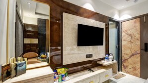 Un baño moderno con un espejo grande, un televisor de pantalla plana, paredes de mármol y un tocador con un recipiente de artículos de aseo.