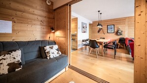 Un interior moderno con una pared de madera, un sofá gris con cojines de cuero de vaca, un comedor con mesa y sillas, y una cocina a la vista.