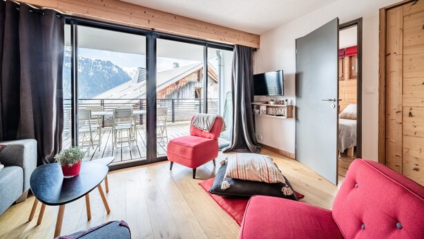 Un salón moderno con una silla roja, un sofá, un televisor y un amplio ventanal corredero que da a un área exterior con vistas a montañas.