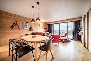 A cozy room with a wooden wall, a round wooden table with chairs, a red sofa, and a large window with curtains.