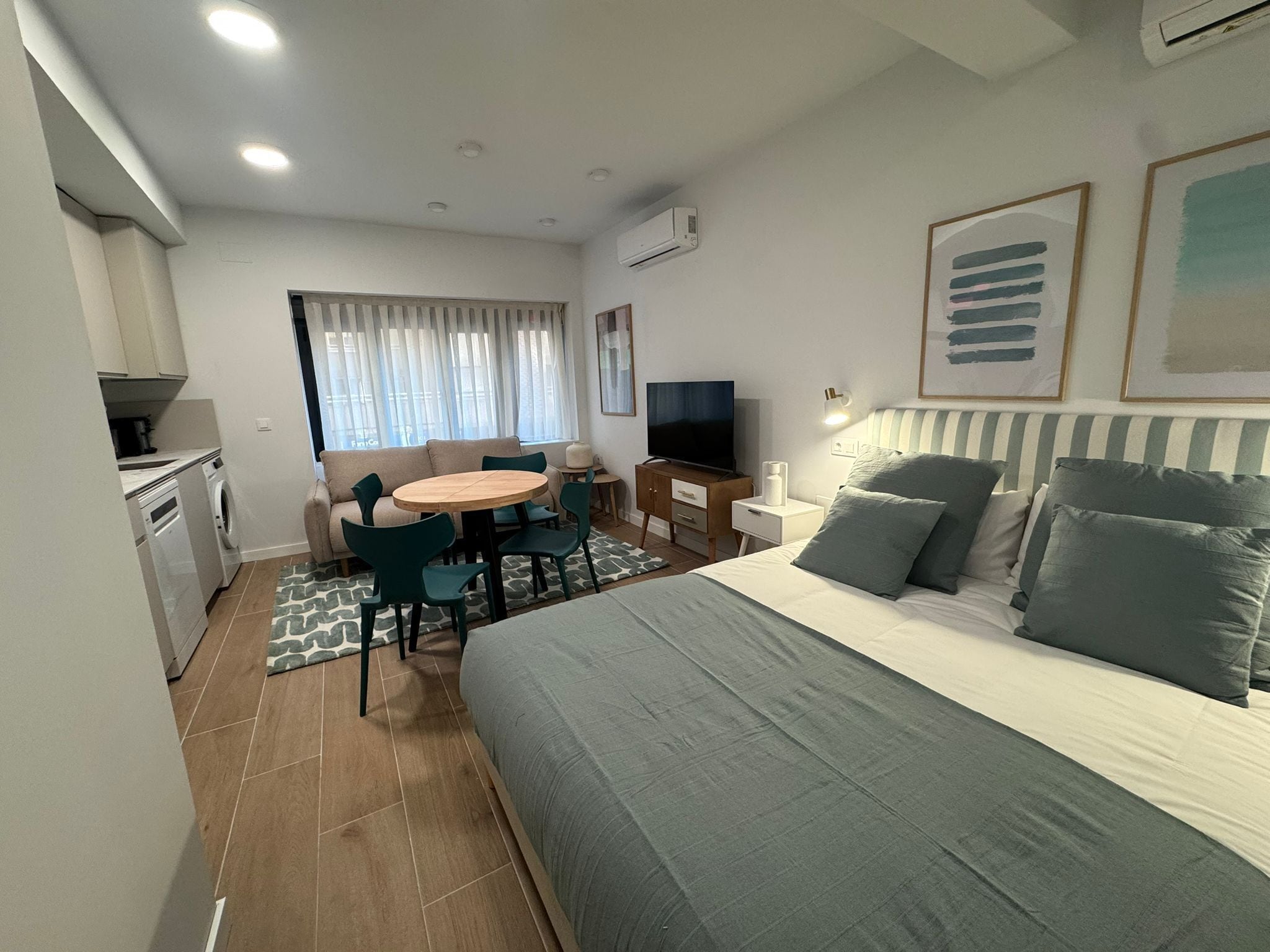 Camera d'albergo compatta con un letto, una TV, un'area pranzo con sedie, una piccola cucina con lavatrice e una finestra con tende.