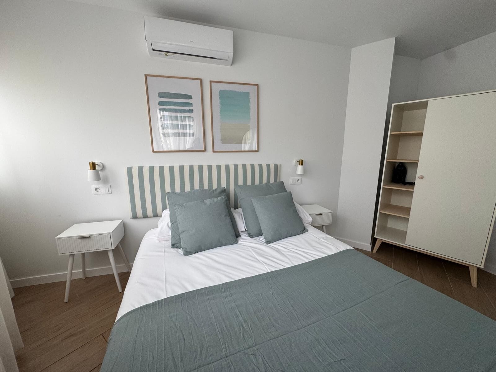 Un dormitorio con cama, mesitas de noche, aire acondicionado, armario y dos cuadros en la pared.
