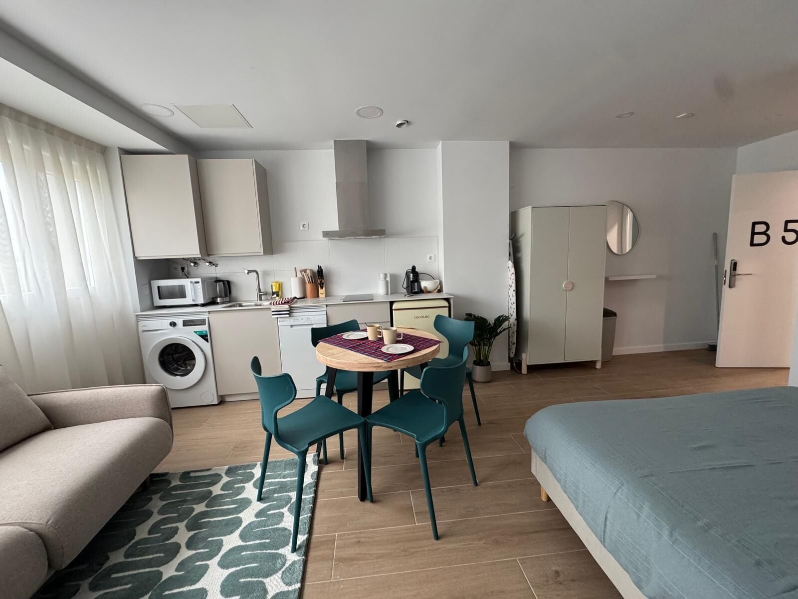 A compact hotel room with a bed, a sofa, a dining table with chairs, a kitchen area, and a wardrobe.