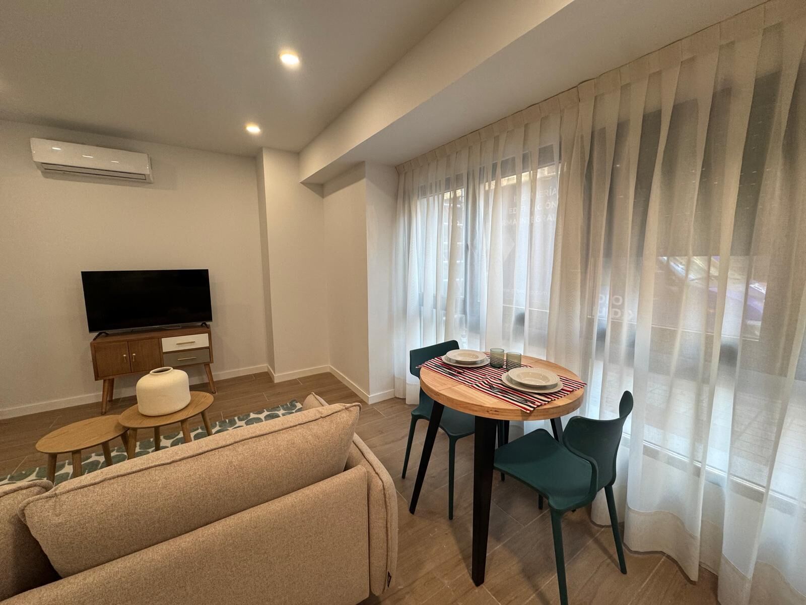 Una sala de estar moderna con una televisión de pantalla plana, un sofá, una mesa de comedor con sillas y cortinas de gasa.