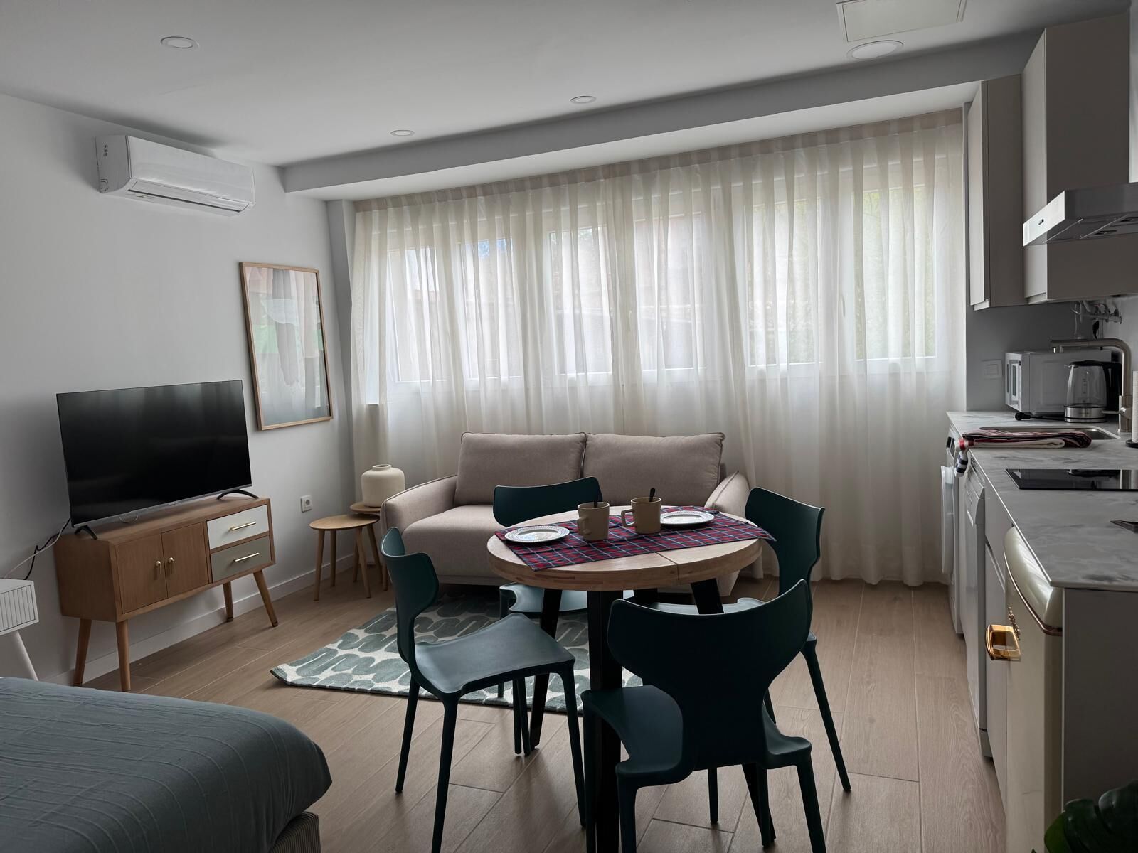 A modern living room with a sofa, TV, dining table, chairs, kitchen area, and large windows with sheer curtains.