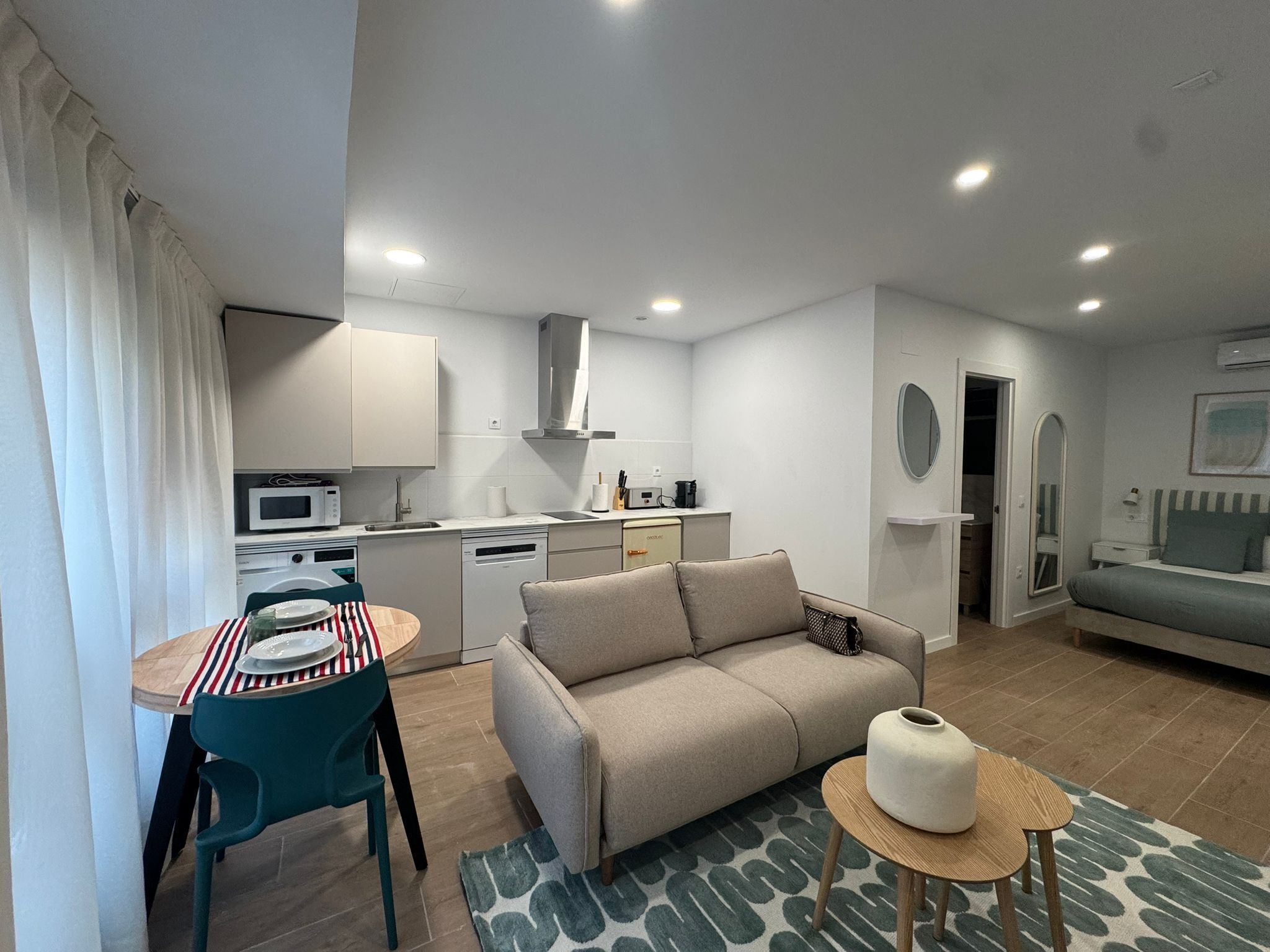 Una sala moderna con sofá, un comedor con mesa y sillas, una cocina equipada y un baño con espejo e inodoro.