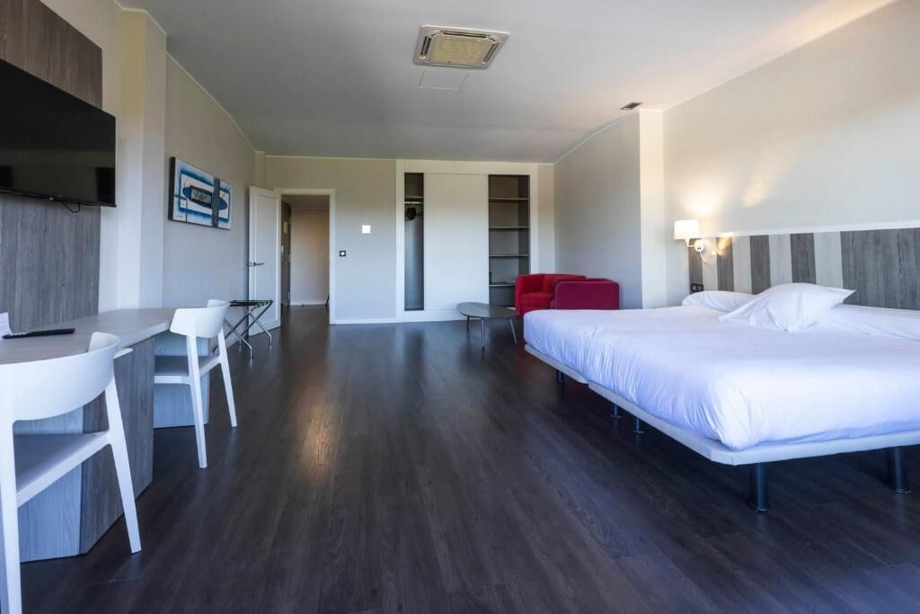 Habitación de hotel moderna con cama, escritorio con sillas, televisor y una silla roja.
