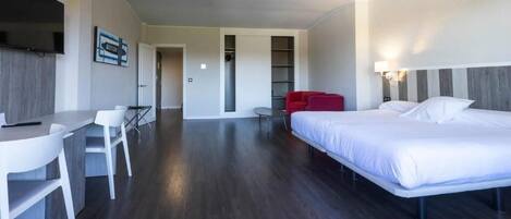 A modern hotel room with a bed, a desk with chairs, a TV, and a red chair.