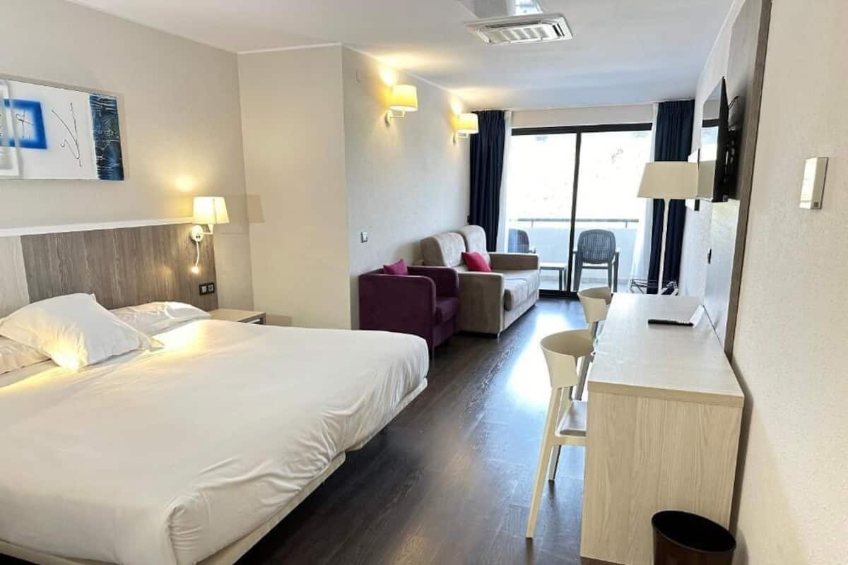 A hotel room with a bed, a desk, a chair, a sofa, and a balcony with a view.