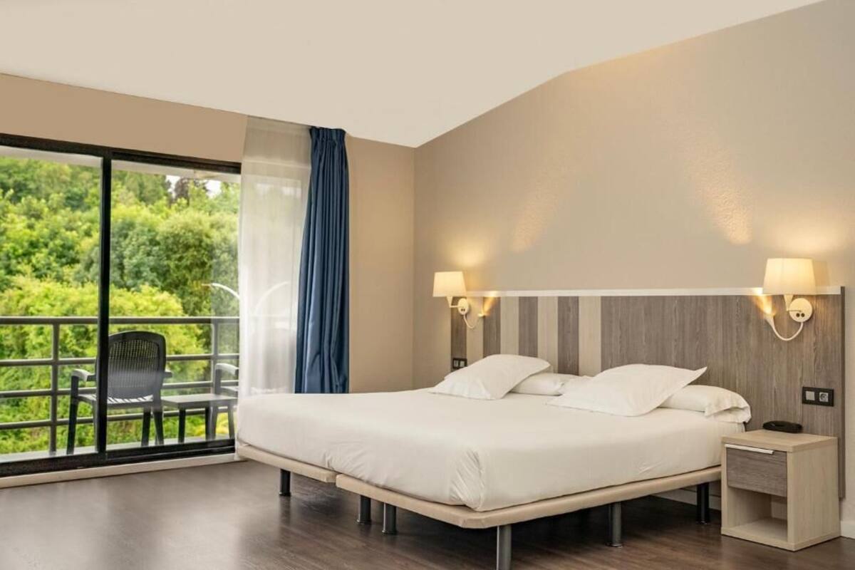 A modern hotel room with a large bed, two bedside lamps, a small bedside table, and a balcony with a chair.
