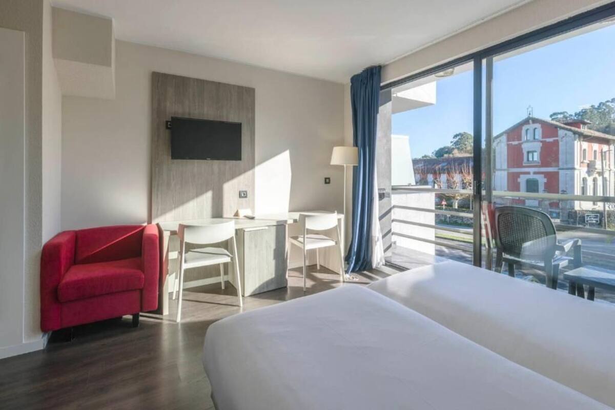 A hotel room with a bed, a desk, a chair, a TV, and a view of buildings outside.