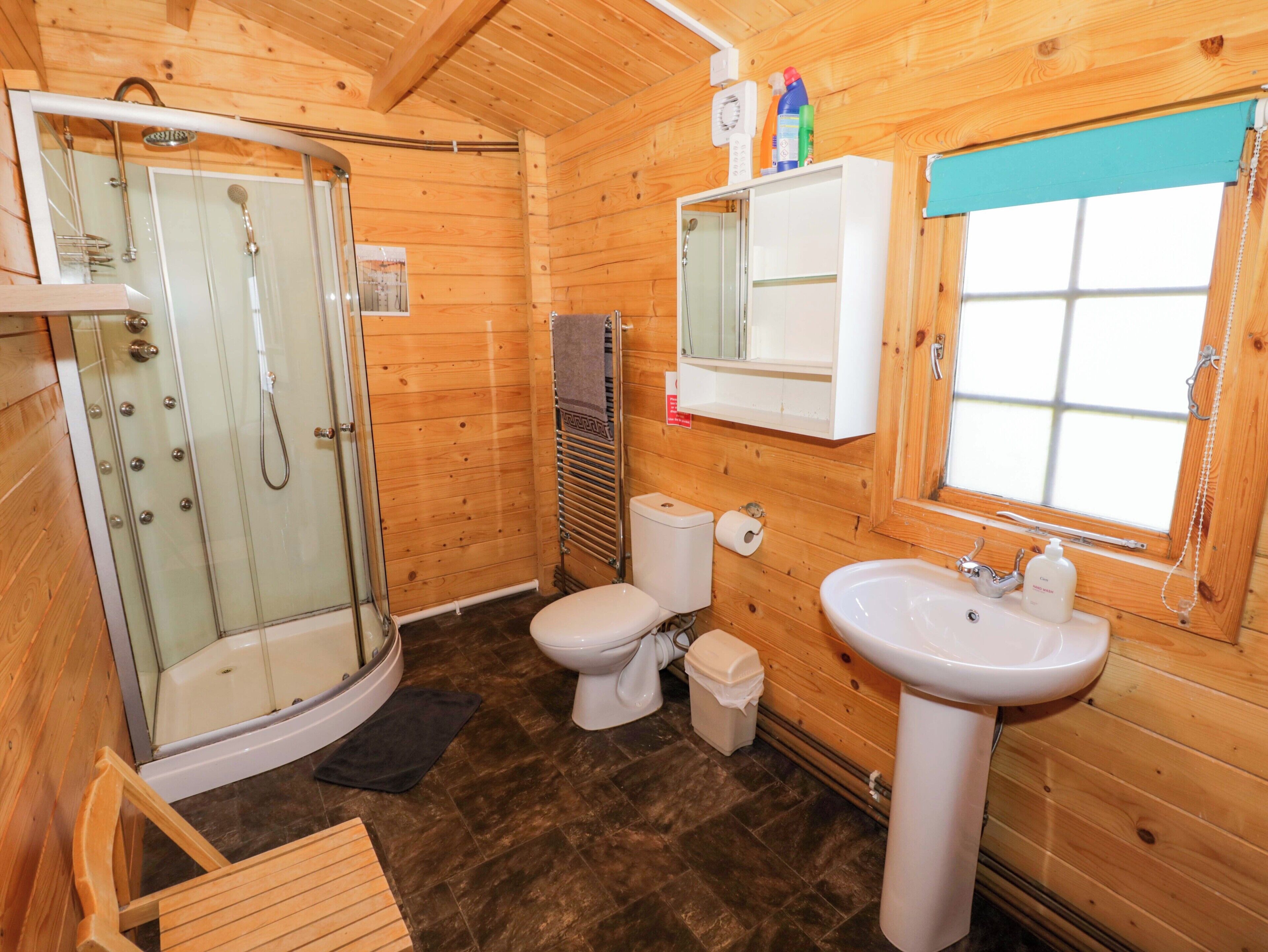 Ein Badezimmer in einer Holzhütte mit Duschkabine aus Glas, Toilette, Waschbecken und Fenster.