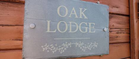 A slate sign with 'OAK LODGE' written on it, mounted on a wooden wall with visible horizontal planks.