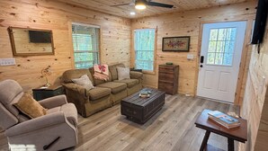 Ein gemütliches Zimmer mit Sofa, Sessel, Holzwänden, Deckenventilator und einer Tür.