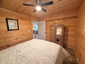A wooden-paneled bedroom with a bed, a dresser, a ceiling fan, and a door leading to another room. - Hot Tub Cabin Retreat • Near Montgomery & Troy, AL (Ramer)