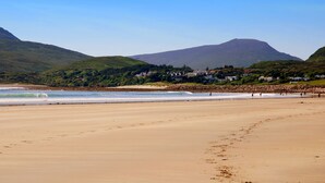 Ein Strand mit einem Berg im Hintergrund, Häuser entlang der Küste und Menschen, die am Sand entlanggehen.