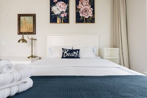 Een netjes opgemaakt bed met een kussen met de tekst 'Beach', een bedlamp en twee bloemrijke schilderijen aan de muur.